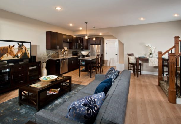 Lofts at Saratoga living room with a grey couch, situated next to a kitchen with stainless steel appliances and brown cabinets.